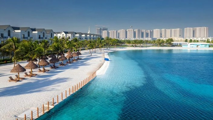 Bãi biển nhân tạo với hàng dù che và ghế nghỉ, nước biển xanh trong và dãy biệt thự ven hồ